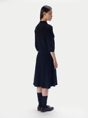 A model shown from the back wearing a black three-quarter sleeve midi dress, black knee-high socks, and black leather shoes against a plain light gray studio background.