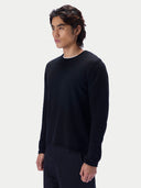 A man shown in three-quarter profile wearing a black crewneck knit sweater with a rolled neckline over a white t-shirt and dark trousers, against a light gray studio background.