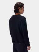 A man shown from the back wearing a black long-sleeve crewneck knit sweater and dark trousers, against a light gray studio background.