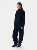 A model wearing a black knit two-piece set consisting of a mock-neck long-sleeve top and matching wide-leg trousers, paired with black leather shoes, against a light gray studio background.
