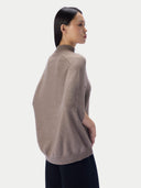 A woman shown from the side profile wearing a taupe knit mock-neck sweater and black pants against a light gray studio background.
