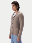 A man shown in three-quarter profile wearing a taupe knit blazer-style cardigan with patch pockets over a white t-shirt and light blue jeans, against a light gray studio background.