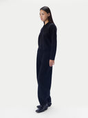 A model stands in profile wearing a dark navy long-sleeve knit jumpsuit with a high mock neck and wide-leg pants, paired with dark leather shoes, against a plain light gray studio background.