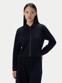 A model wearing a black long-sleeve knit top with a unique collar and front zipper, paired with black trousers, against a light gray studio background.