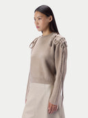 A woman wearing a beige ribbed knit sweater with knotted decorative details on the shoulders and sleeves, paired with a light beige skirt, photographed against a plain light gray studio background.