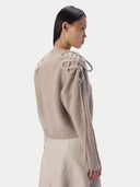 A model shown from the back wearing a ribbed knit sweater in a light taupe-beige color, featuring knotted decorative details on the shoulders and long tie-like strands hanging from the sleeves, paired with light beige trousers against a plain light gray background.