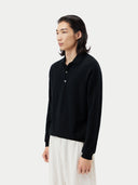 close up side view of a person wearing a black cashmere polo sweater with a collar and white pants, standing against a plain background.  #color_black