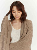 A front view of a person with wavy brown hair looking downward, wearing a taupe cable-knit cardigan over a white textured top against a white background. #color_taupe