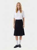 A woman stands against a light gray studio background wearing a white short-sleeve crewneck T-shirt, a black A-line midi skirt, white knee-high socks, and black leather loafers.