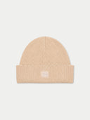 A light beige ribbed knit beanie hat with a folded cuff and a small white embroidered logo in the center, shown against a light gray background.