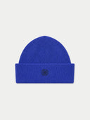 A bright royal blue ribbed knit beanie hat with a folded cuff and a small black embroidered geometric flower-like logo in the center, shown on a light gray background.