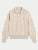 A light beige knit polo sweater with a three-button placket and a small embroidered circular logo on the left chest, displayed on a clean off-white background.