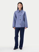 A model stands against a light gray studio background wearing a light periwinkle blue cropped jacket with dark buttons and dark navy wide-leg trousers.