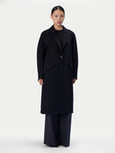 A model stands against a plain light gray studio background wearing a long black coat over a black top and dark wide-leg trousers.