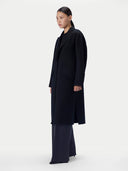 A model in side profile wearing a long black coat over a black outfit, standing against a plain light gray studio background.