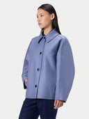 A woman wearing a light blue-gray structured coat with dark buttons, paired with dark navy trousers, photographed against a clean light gray studio background.