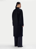 A model shown from the back wearing a long black coat and wide-leg trousers against a plain light gray studio background.