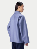 A model shown from the back wearing a light periwinkle blue oversized blazer with dark navy trousers, set against a plain light gray studio background.