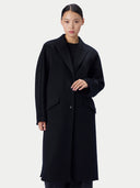 A model wearing a long black wool-blend coat with a single button closure, large notched lapels, and flap pockets, photographed against a light gray studio background.