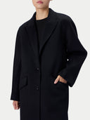A model wearing a black wool-blend coat with notched lapels, three front buttons, and flap pockets, paired with a black top against a light gray studio background.