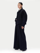 A model is shown in profile wearing a dark navy blue jacket and matching wide-leg skirt against a plain light gray studio background.