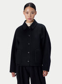 A model wearing a black button-up jacket with a pointed collar and large front patch pockets, paired with black trousers and a cream shirt, shot against a light gray studio background.