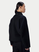 A model shown from the back wearing a black cropped jacket with a high mock neck collar, photographed against a solid light gray background.