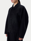 A person wearing a black button-front jacket with a collar and large front patch pockets, photographed against a plain light gray studio background.