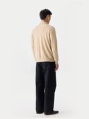 back view of a man wearing a beige sweater with a collar and black pants on a white background.