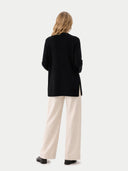 back view of a woman wearing a black cardigan and beige pants on a white background.