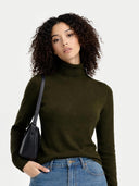 Woman wearing a dark green turtleneck sweater and blue jeans, holding a black handbag on a light gray background. #color_avocado