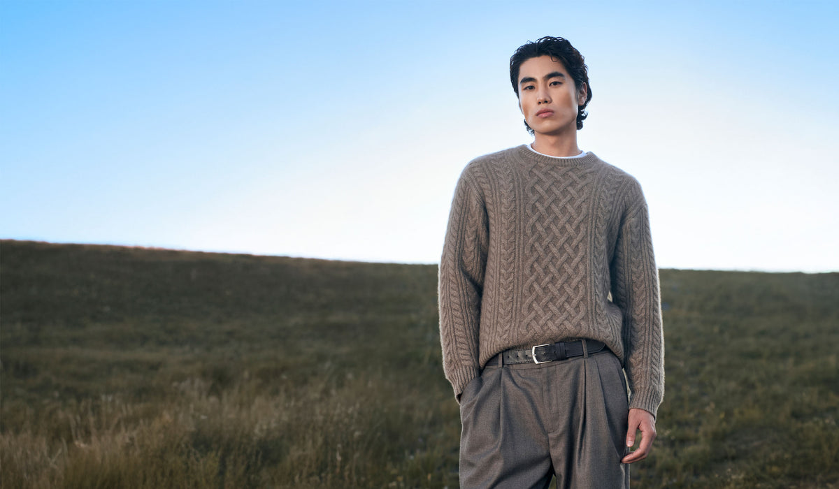Man in a taupe cable-knit cashmere sweater and gray trousers standing on a grassy hillside under a blue sky
