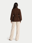 back view of a woman wearing a brown coat and beige pants on a white background