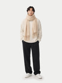 front view of a man wearing a white sweater, black pants and a beige scarf with fringes on a white background.