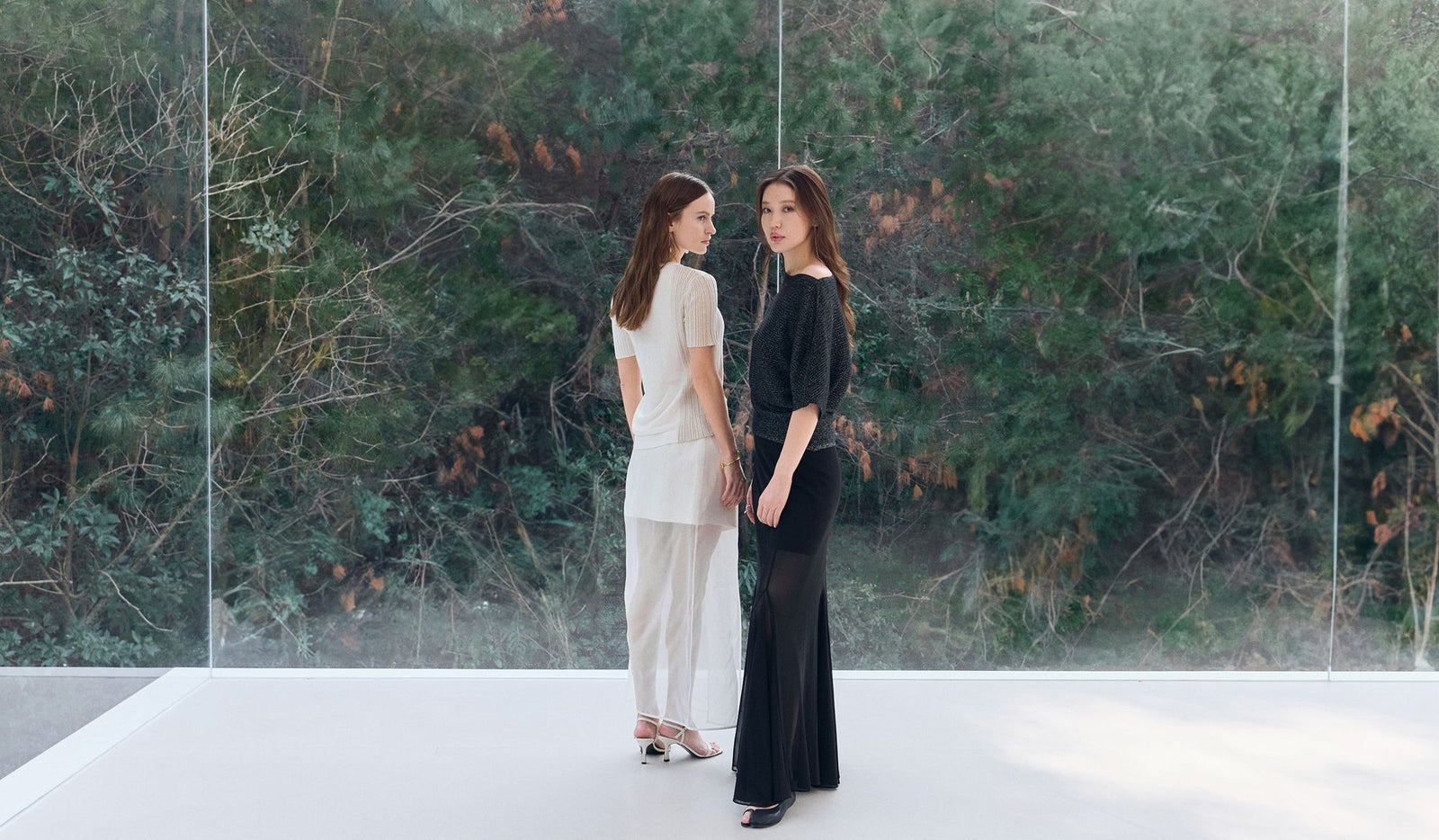 Two women standing on a white platform with a reflective surface and greenery in the background