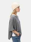 Woman wearing a gray poncho, white beanie, and blue jeans on a light background. #color_off-white