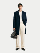 Man wearing a navy coat, white shirt, beige pants, and holding a brown leather bag on a white background #color_navy