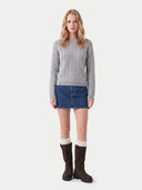 A model stands against a light gray studio background wearing a light gray cable-knit sweater, a blue denim mini skirt, cream ribbed socks, and dark brown knee-high boots.