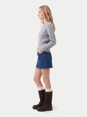A woman shown in profile wearing a light gray cable-knit sweater, a blue denim mini skirt, cream socks, and dark brown mid-calf boots, photographed against a plain light gray studio background.