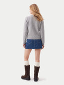A woman shown from the back wearing a light gray cable-knit sweater, a blue denim mini skirt, cream ribbed socks, and dark brown boots, against a plain light gray studio background.