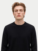 A man with light brown hair wearing a black long-sleeve crewneck sweater, photographed against a solid light gray background.