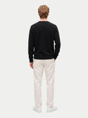 A man viewed from behind wearing a black long-sleeve crewneck sweater, off-white trousers, and white sneakers, standing against a light gray studio background.