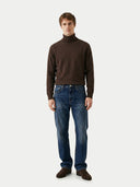 Man wearing a brown turtleneck sweater and blue jeans on a white background.
