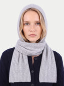 A woman wearing a light gray ribbed knit hooded scarf and matching neck warmer over a dark navy cardigan, shot against a plain light gray background.
