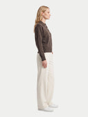A woman stands in profile wearing a dark brown knit button-front cardigan, off-white wide-leg trousers, and white sneakers against a plain light gray studio background.