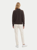 A woman viewed from the back wearing a dark brown long-sleeve knit top, off-white trousers, and white sneakers against a plain light gray studio background.