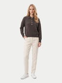 A woman stands against a light gray studio background wearing a dark brown long-sleeve polo sweater with a buttoned collar, off-white trousers, and white sneakers.