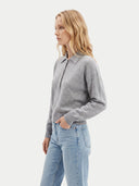 A woman wearing a light gray long-sleeve polo sweater and light blue jeans, shown in profile against a plain light gray studio background.