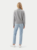 A woman viewed from the back wearing a light gray knit mock-neck sweater, light-wash blue jeans, and white sneakers against a plain light gray studio background.