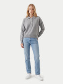A woman modeling a gray long-sleeve knit polo sweater with light blue jeans and white sneakers, standing against a plain light gray studio background.